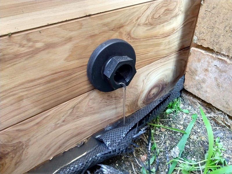 A plastic drain outlet in the side of a wooden garden bed with a small amount of water dripping from it.